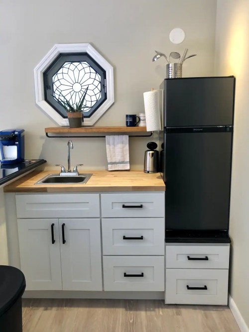 A modern kitchen corner featuring a sink, black fridge, wooden shelf, and a decorative octagonal window.