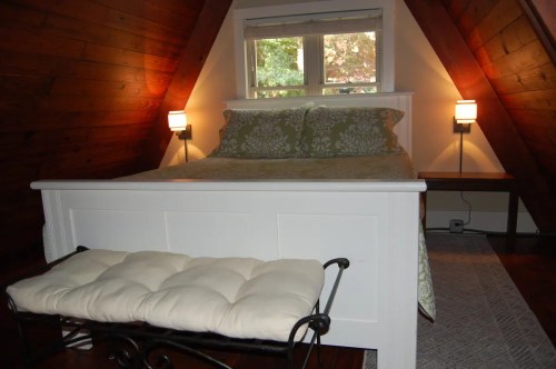 Cozy attic bedroom with a white bed, green bedding, and wooden walls, featuring a bench and soft lighting.