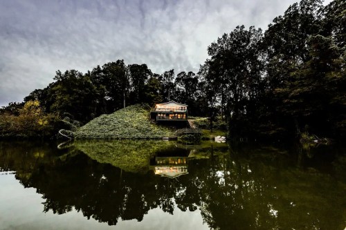 A modern house on a hill by a calm lake, surrounded by trees, reflecting in the water under a cloudy sky.