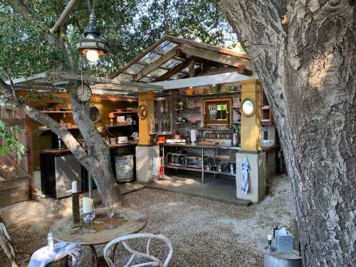 A cozy outdoor kitchen surrounded by trees, featuring a rustic design and a gravel seating area.