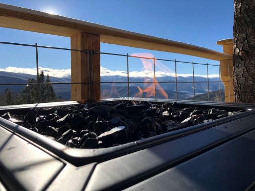 A close-up of a fire pit with flames, set against a scenic mountain backdrop and clear blue sky.