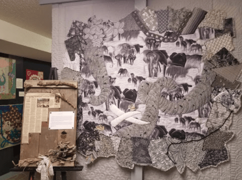 A quilt featuring elephants, surrounded by various fabric patterns, displayed alongside an informational board.