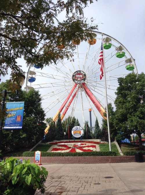 A colorful Ferris wheel at a park in Denver, surrounded by greenery and flowers, with an American flag nearby.