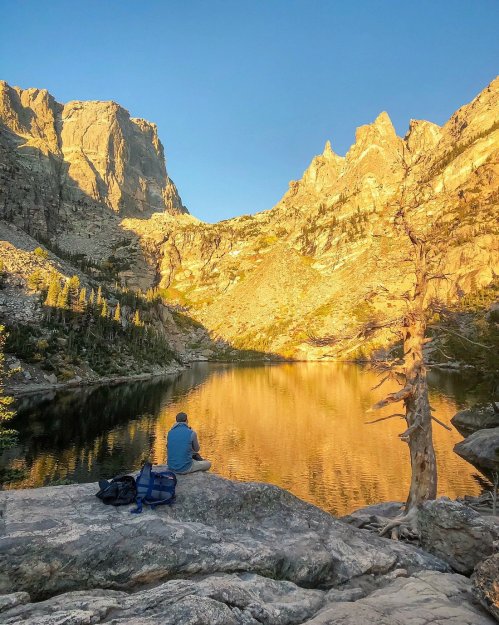 A person sits on a rock by a serene lake, surrounded by mountains reflecting golden sunlight at sunset.