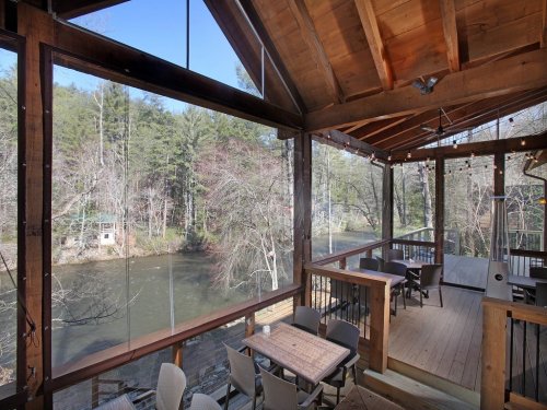 A cozy wooden deck overlooking a river, surrounded by trees, with outdoor seating and a clear blue sky.
