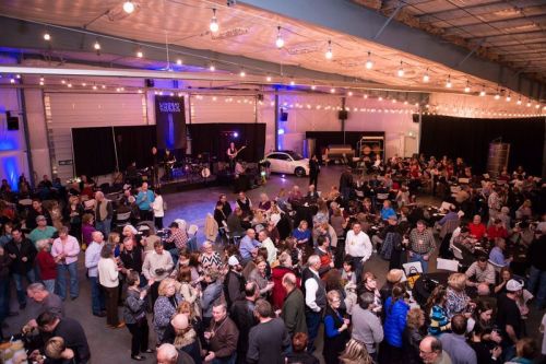 A crowded indoor event with people socializing, a band performing on stage, and festive lighting.