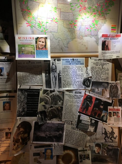 A bulletin board covered with missing person posters, newspaper clippings, and a map of Texas marked with locations.