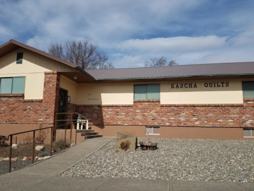 Exterior of a brick building with a sign that reads "Kascha Quilts," featuring a sloped roof and a gravel pathway.