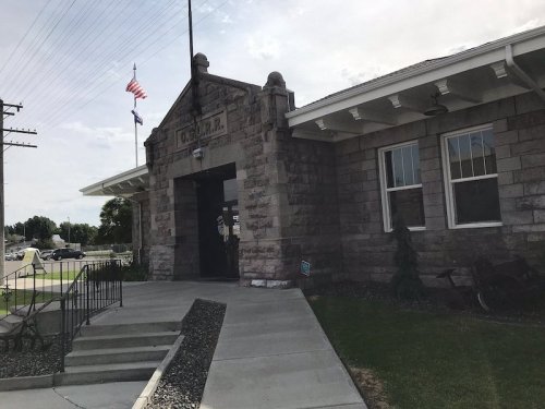 Historic stone train station with flags, steps, and a grassy area in front.