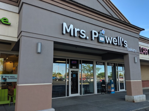Exterior of Mrs. Powell's Bakery, featuring a sign and large windows, with nearby shops visible in the background.