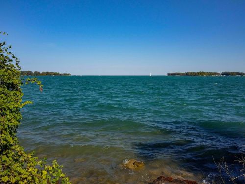 A serene view of a blue lake with gentle waves, framed by green foliage and distant islands under a clear blue sky.