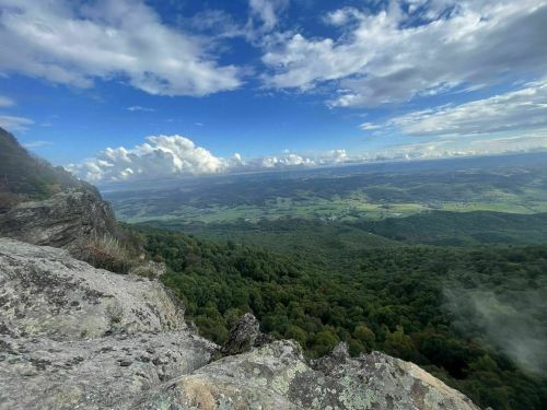 A panoramic view from a rocky ledge overlooking lush green valleys and distant hills under a partly cloudy sky.