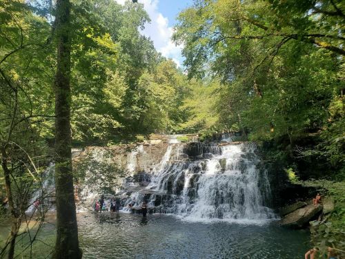 A serene waterfall surrounded by lush greenery, with people enjoying the water at its base on a sunny day.