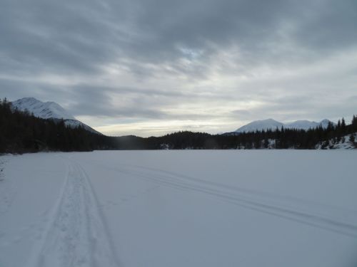 A snowy landscape with a frozen lake, surrounded by mountains and overcast skies. Snowmobile tracks lead into the distance.