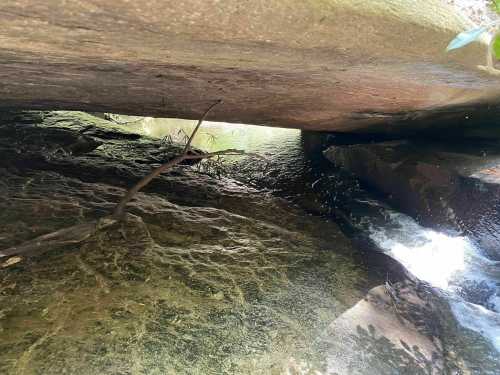 A narrow passage under a large rock, with water flowing over smooth stones and greenery in the background.