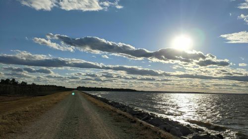A scenic view of a gravel path along a shimmering lake, with clouds and a bright sun in the sky.