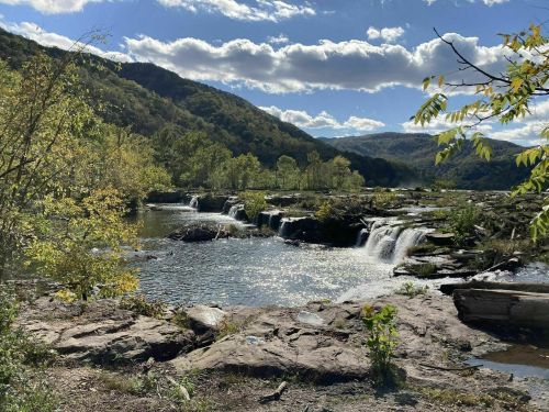 A serene landscape featuring a waterfall cascading into a calm river, surrounded by lush greenery and mountains under a blue sky.