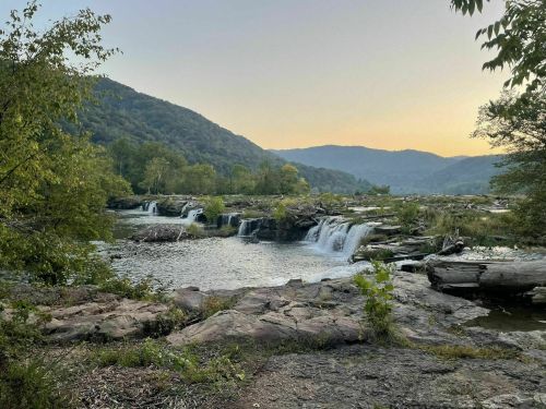 A serene landscape featuring waterfalls cascading over rocks, surrounded by lush greenery and distant mountains at sunset.