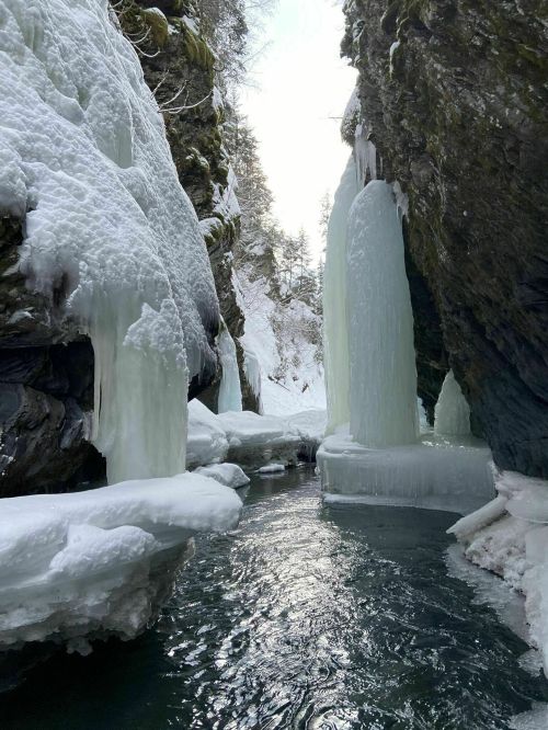A serene winter scene of icy cliffs and frozen waterfalls beside a flowing river in a narrow canyon.