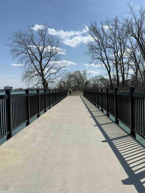 A concrete walkway extends along a river, bordered by black railings and trees under a clear blue sky.