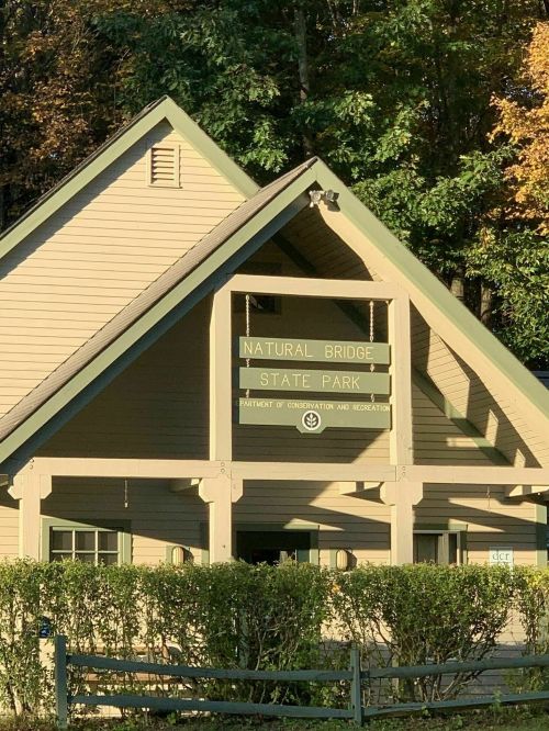 A wooden building with a triangular roof, featuring a sign that reads "Natural Bridge State Park" surrounded by greenery.