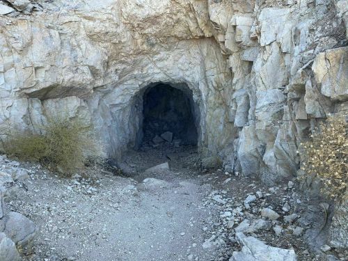 A rocky cave entrance surrounded by dry vegetation and gravel, leading into darkness.