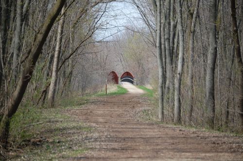 A dirt path leads through trees to a covered bridge in the distance, surrounded by early spring foliage.