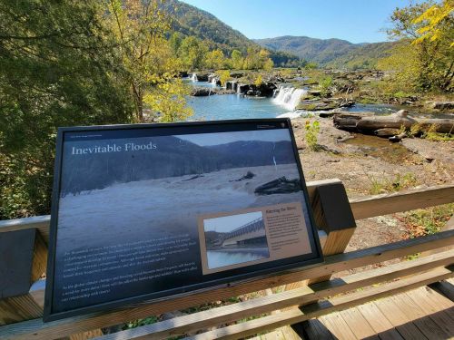 A sign about inevitable floods near a river with a waterfall and mountains in the background on a sunny day.