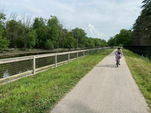 A child rides a bike along a paved path beside a river, surrounded by trees and greenery on a sunny day.
