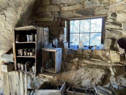A rustic stone wall interior with a wooden shelf holding jars and pots, and a window letting in natural light.