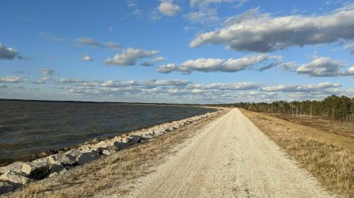 A gravel path alongside a lake, with clouds in a blue sky and trees in the background.
