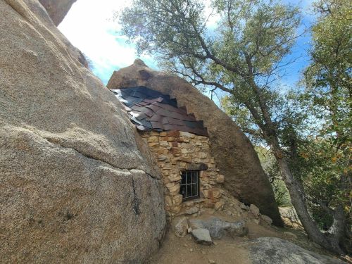 A small stone cabin nestled between large boulders, surrounded by trees and a bright blue sky.