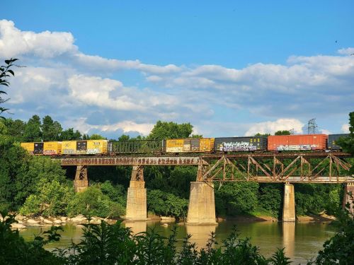 A freight train crosses a metal bridge over a river, surrounded by lush green trees and a blue sky with clouds.