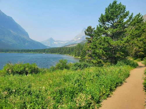 A scenic view of a lake surrounded by mountains and lush greenery, with a walking path along the water's edge.