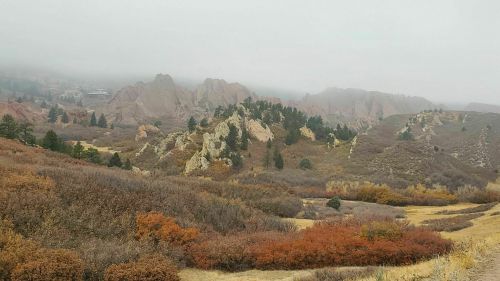 Foggy landscape featuring rocky hills, evergreen trees, and autumn foliage in shades of orange and brown.