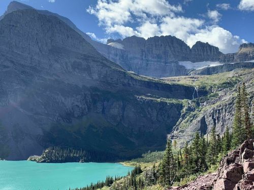 A stunning landscape featuring a turquoise lake surrounded by mountains and lush greenery under a partly cloudy sky.