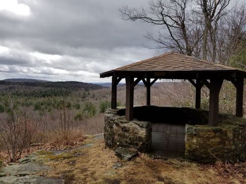 A stone gazebo overlooks a cloudy landscape of trees and hills, with a rocky foreground and bare branches.