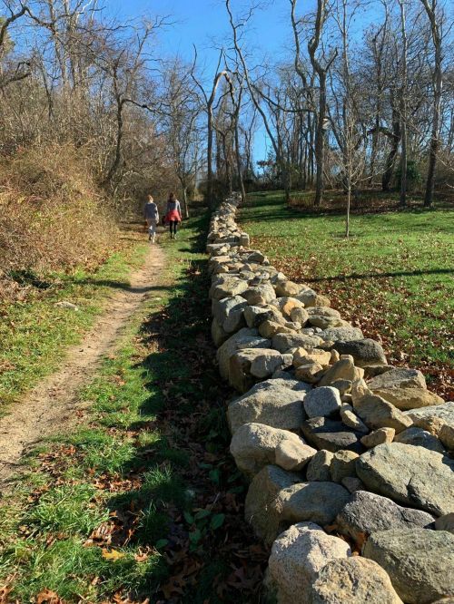 Two people walk along a dirt path beside a stone wall, surrounded by trees and autumn foliage under a clear blue sky.