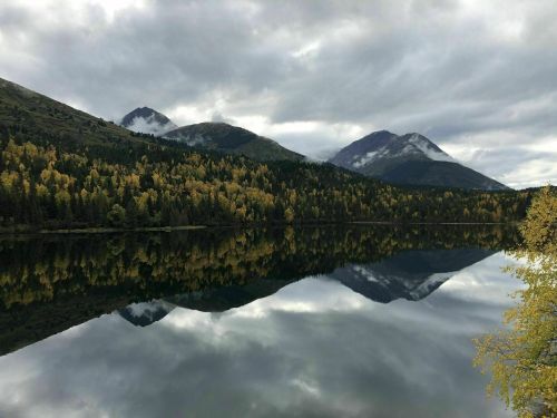 A serene lake reflects mountains and autumn foliage under a cloudy sky.