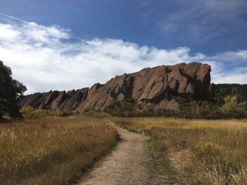A dirt path leads through tall grass towards a rocky formation under a blue sky with scattered clouds.