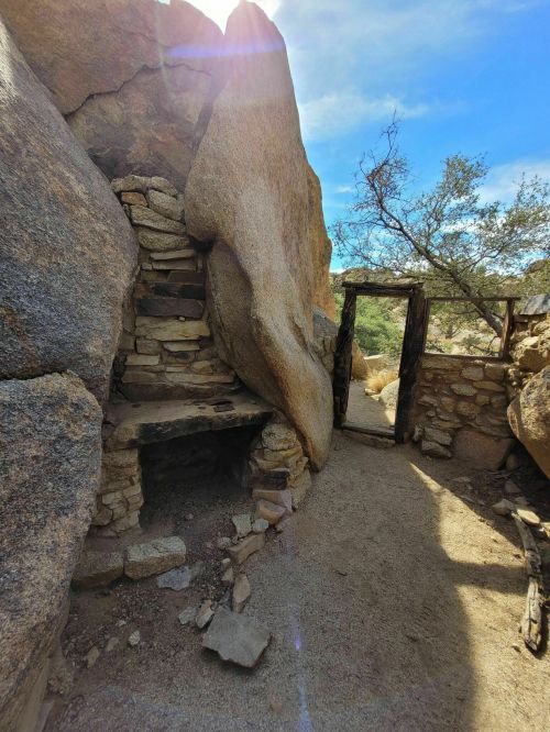 A rocky landscape featuring a narrow passageway with stacked stones and a wooden door, surrounded by desert vegetation.