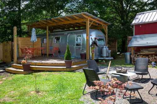 A silver Airstream trailer under a wooden pergola, surrounded by chairs and a small fire pit in a green yard.