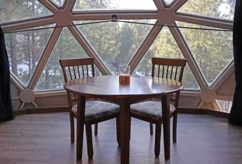 A wooden table with two chairs, set against a large geometric window overlooking a forested landscape.