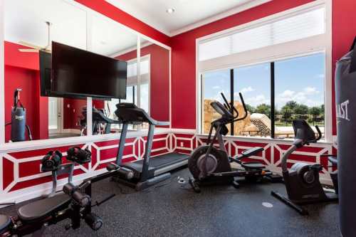 Home gym with red walls, treadmill, elliptical, weights, and a large mirror reflecting a sunny outdoor view.