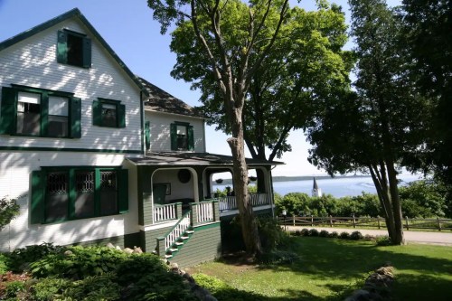 A charming white house with green shutters, surrounded by trees, overlooking a scenic view of water and distant hills.