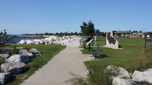 A scenic pathway along a waterfront, lined with rocks, benches, and informational signs under a clear blue sky.