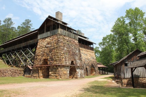 Historic stone structure with wooden upper levels, surrounded by trees and a dirt path, showcasing rustic architecture.