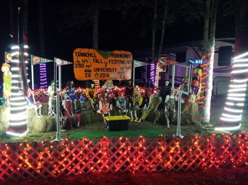 A festive Halloween display with skeletons, hay bales, and colorful lights, set in a dark outdoor area.