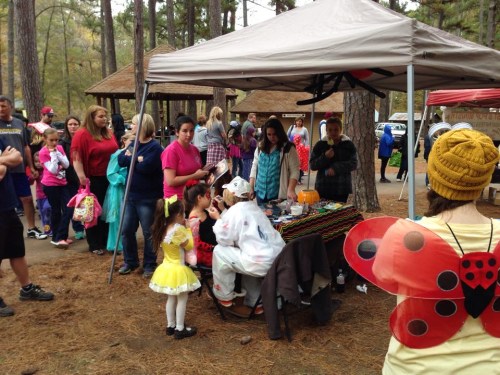 A crowd gathers around a table under a tent in a wooded area, with children in costumes and adults chatting.