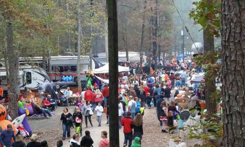 A bustling campground filled with people, tents, and decorations, surrounded by trees in autumn.